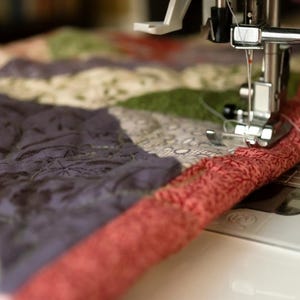 May include: A sewing machine needle and presser foot stitching a quilt with a red patterned fabric border. The quilt features triangular sections of dark purple, green, and cream-colored fabric with intricate floral and leaf patterns.