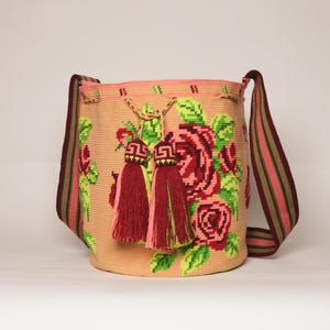 Peut inclure: Sac à bandoulière beige tissé avec un motif floral de roses rouges et vertes. Le sac a une fermeture à cordon et deux pompons décoratifs. La bandoulière est rayée de rouge, vert et marron. Le sac mesure environ 30 cm de large et 25 cm de haut.
