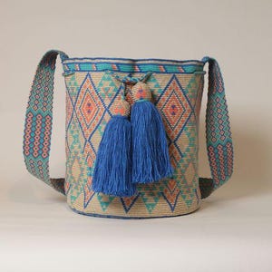 May include: A woven shoulder bag with a geometric diamond pattern in shades of blue, peach, and tan. The bag has a wide strap and is adorned with two large blue tassels. The bag's dimensions are not visible.