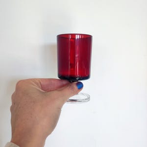 May include: A vibrant red glass with a clear stem. The glass has a cylindrical shape and a wide opening. The stem is short and the base is round. The glass is held against a white background.