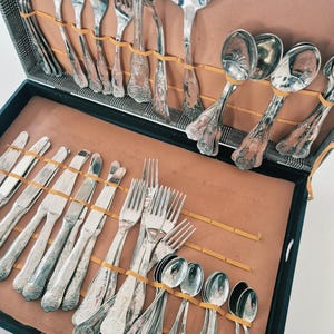 Può includere: Un set di posate color argento disposto in una custodia nera con interno color cuoio. La collezione comprende varie forchette, coltelli e cucchiai, ognuno caratterizzato da dettagli in rilievo sui manici, fissati da elastici gialli.
