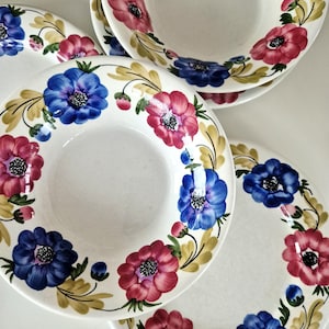 May include: A set of white ceramic bowls and plates featuring a hand-painted floral pattern. The design includes vibrant blue and pink anemone-style flowers with dark centers, accented by soft green leaves and small buds arranged around the rims.