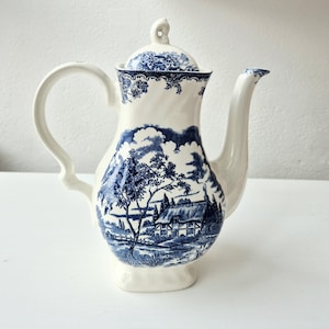 May include: A white ceramic coffee pot with a blue and white landscape design. The pot has a curved handle, a long spout, and a lid with a small knob. The landscape scene includes trees, a house, and clouds.