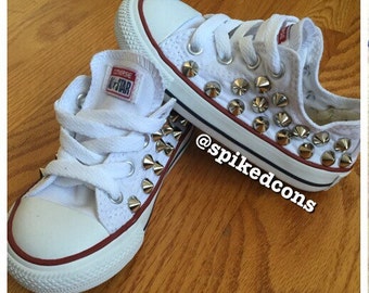 May include: White Converse All Star sneakers customized with silver cone studs. The shoes have white laces, a red stripe around the sole, and a white rubber toe cap. The text "spikedcons" is visible on the side.