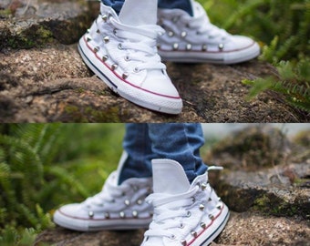 May include: White high-top sneakers with silver studs on the sides and laces. The sneakers are worn with blue jeans.