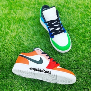 May include: Two pairs of custom-painted sneakers with different color combinations. One pair is orange, red, and black, and the other is green, blue, and white. The sneakers have the text "@spikedcons" on the side.
