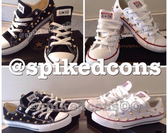 May include: Black and white Converse All Star sneakers with gold-colored studs. The shoes have white laces and soles, with the All Star logo on the tongue. White Converse All Star sneakers with gold studs are also shown.