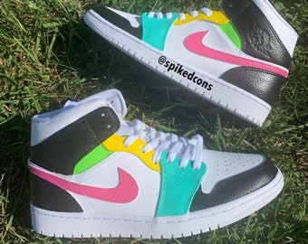Könnte beinhalten: Ein Paar weiße, schwarze, rosa, gelbe und türkisfarbene High-Top-Sneaker mit einem Nike-Swoosh-Logo. Die Sneaker befinden sich auf einer grünen Grasfläche. @spikedcons