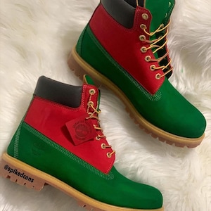 Può includere: Un paio di stivali verdi, rossi e neri con occhielli e lacci dorati. Gli stivali hanno una suola marrone e un'etichetta con un logo. Gli stivali sono su una superficie bianca e soffice. Il testo "@spikedcons" è visibile sulla suola.