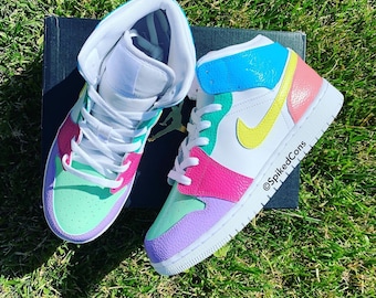 May include: A pair of colorful high-top sneakers with a white base and accents in pastel shades of pink, green, yellow, and blue. The shoes are on a black box, set against a backdrop of green grass. The text "@SpikedCons" is visible.