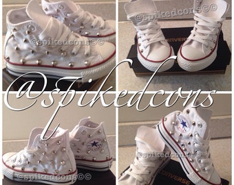 May include: White high-top sneakers with silver studs. The shoes have white laces, a red and blue stripe on the sole, and a Converse logo on the side. The shoes are displayed on a black box.
