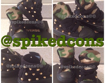 May include: Black high-top sneakers with gold-colored studs. The sneakers have a camouflage pattern on the tongue and collar. The word "CONVERSE" is visible on the box.