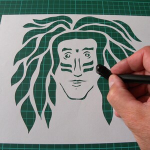 May include: A white paper stencil with a detailed cutout of a person's face with long hair. The person has a tribal-style design painted on their face.