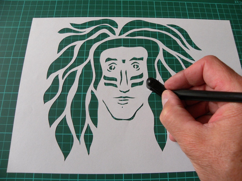 May include: A white paper stencil with a detailed cutout of a person's face with long hair. The person has a tribal-style design painted on their face.