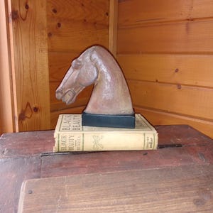 May include: A brown horse head sculpture on a black base, resting on two copies of the book "Black Beauty" by Anna Sewell. The books are tan with black text. The sculpture is set on a wooden table.