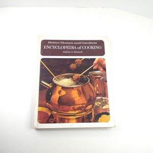 May include: A Better Homes and Gardens Encyclopedia of Cooking book with a white cover and a dark brown rectangular header. The cover features the text "Better Homes and Gardens ENCYCLOPEDIA of COOKING Abalone to Bannock" above a photo of a copper fondue pot.