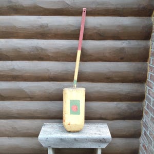May include: A vintage, weathered wooden buoy with a long handle. The buoy is painted yellow with a green and red design. The handle is painted in sections of red, yellow, and green. The buoy is displayed on a small, rustic wooden table.