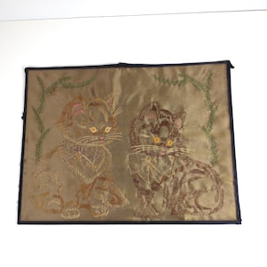 May include: Embroidered artwork featuring two kittens with golden eyes and detailed fur patterns. The kittens are set against a gold background with green embroidered foliage. The artwork is framed with a black border.