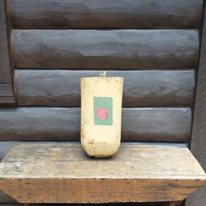 May include: A weathered, cylindrical wooden buoy painted in cream with a green square and a red circle. The buoy sits on a rustic wooden shelf against a backdrop of a log cabin wall. The buoy has a small loop at the top.