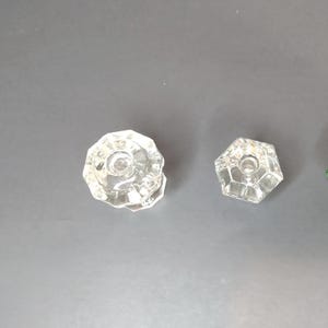 May include: Two clear glass cabinet knobs with faceted surfaces. The larger knob on the left features a multi-sided circular design, while the smaller knob on the right has a distinct hexagonal shape. Both knobs have a central hole for mounting.
