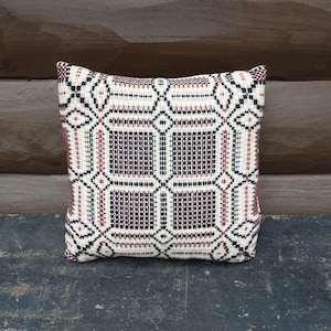 May include: Square decorative pillow with a geometric pattern in cream, black, and burgundy. The pillow has a woven texture and is placed on a dark, weathered surface. The background is a wooden wall.