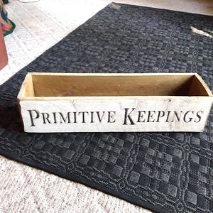 May include: A rectangular wooden box with the words "PRIMITIVE KEEPINGS" in black lettering. The box is painted white with a distressed finish, and the interior is a natural wood color. The box is on a dark gray patterned rug.