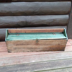 May include: A vintage wooden toolbox with a hinged lid and a handle. The box is painted in shades of brown and teal, with metal hardware. The toolbox is likely used for storing tools and other small items.