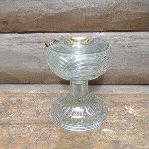 May include: A clear glass oil lamp base featuring a rounded reservoir with embossed swirling wave patterns and a wide, fluted pedestal foot. The top includes a threaded brass collar and a small, circular metal filler cap on the side.