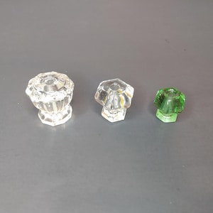 May include: Three glass cabinet knobs. Two are clear, faceted, and have a pedestal base. The third knob is green, also faceted, and has a hexagonal base. All three knobs are on a gray surface.