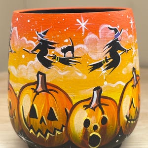 Puede incluir: Una taza de cerámica naranja pintada a mano con temática de Halloween. La taza presenta dos brujas volando en escobas con gatos negros y tres calabazas talladas en forma de jack-o'-lantern.