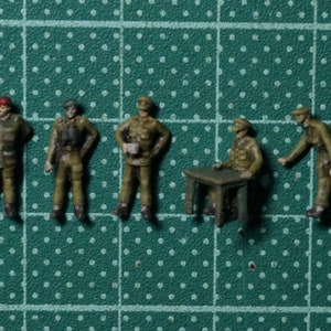 以下が含まれることがあります： 迷彩服を着た兵士のミニチュアフィギュア5体。フィギュアは、小さな木製のテーブルの周りに立ったり座ったりしています。フィギュアはリアルなディテールでペイントされています。