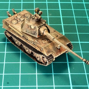 Puede incluir: Un modelo detallado de un tanque alemán, pintado con un patrón de camuflaje marrón, verde y beige. El tanque tiene un cañón largo y una torreta con una escotilla abierta. El modelo está sentado en una estera de corte verde.