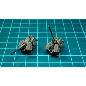 Puede incluir: Dos modelos en miniatura, detallados, pintados, de metal de cañones antiaéreos. Los cañones están sobre un fondo de rejilla verde. Los cañones están pintados en un patrón de camuflaje marrón, verde y negro.