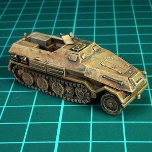Peut inclure: Maquette miniature d'un véhicule militaire, de couleur beige et marron, avec des chenilles et des roues visibles. Le modèle présente un design détaillé, avec un petit canon sur le dessus. Le véhicule est posé sur un fond quadrillé vert.