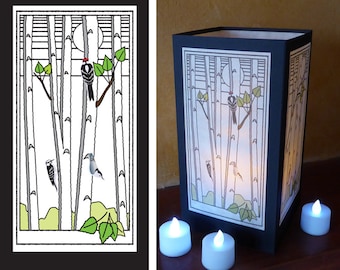 May include: A black and white paper lantern with a design of a moon, trees, and birds. The lantern is lit from the inside, creating a warm glow.