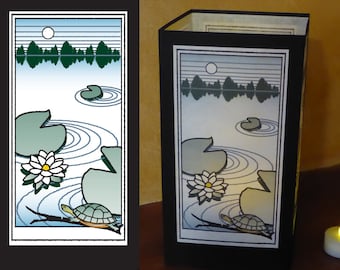 May include: A black lantern with a white and blue illustration of a turtle swimming in a pond with lily pads and a full moon. The illustration is framed in black.