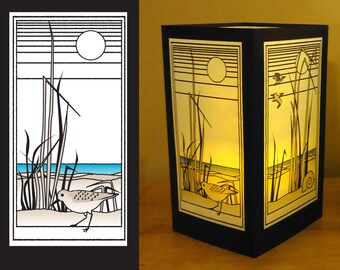 May include: A black framed lamp with a white and yellow design of a bird, grass, and a moon on a beach. The lamp is lit and the light shines through the design.