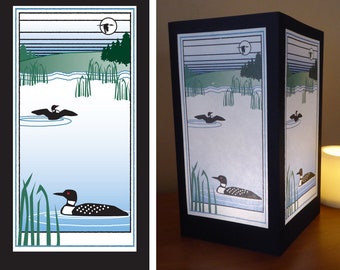 May include: A black and white illustration of a loon on a lake with reeds and a moon in the sky. The illustration is printed on a white paper lampshade with a black frame.