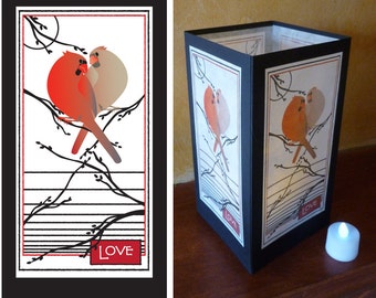 May include: A black paper lantern with a red and beige bird illustration on a white background. The illustration includes branches and the word "LOVE" in a red box. A small, white, battery-operated candle sits nearby.