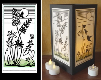 May include: A black silhouette of flowers and a hummingbird with a moon and stripes in the background. The image is on a white background and is inside a black frame. The image is lit from behind with a warm light.