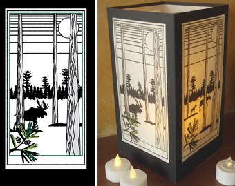 May include: A black and white paper lantern with a silhouette of a moose and trees in a forest scene. The lantern is lit from the inside, casting a warm glow.