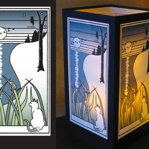 May include: A white cat sits in tall green grass, looking at a small brown frog perched on a blade of grass. The scene is illuminated by a full moon and stars in a blue sky. The image is framed in black and is part of a paper lantern.