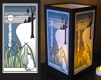 May include: A white cat sits in tall green grass, looking at a small brown frog perched on a blade of grass. The scene is illuminated by a full moon and stars in a blue sky. The image is framed in black and is part of a paper lantern.