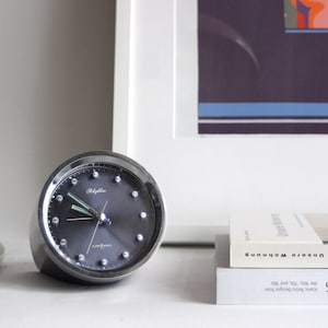 Puede incluir: Un reloj despertador plateado con una esfera negra y agujas blancas. El reloj tiene un borde plateado y una base plateada. El reloj está sobre una superficie blanca.