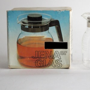 May include: A clear glass teapot with a brown handle and a black lid. The teapot is sitting on a white surface. The box in the background is labeled "JENAer GLAS".