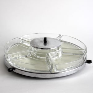 Puede incluir: Una bandeja de servir giratoria plateada con seis cuencos de vidrio transparente y una tapa. La bandeja tiene dos asas negras en el lateral.