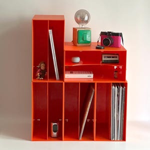 Puede incluir: Un mueble de almacenamiento modular de color naranja brillante con múltiples compartimentos. Contiene una cámara rosa, una pequeña lámpara verde y de madera, una figura de mono y discos de vinilo. Una bombilla se encuentra encima de la lámpara.