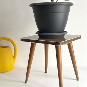 May include: A black plastic plant pot with a matching saucer sits atop a small, square table. The table has a brown surface with a white edge, supported by four wooden legs with brass-colored tips. A yellow watering can is visible in the background.