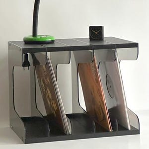 以下が含まれることがあります： 3つのコンパートメントにレコードを収納する黒いレコード収納ユニット。緑色のデスクランプと小さな黒い正方形の時計が上にあります。ランプはフレキシブルネックと緑色のシェードが付いています。