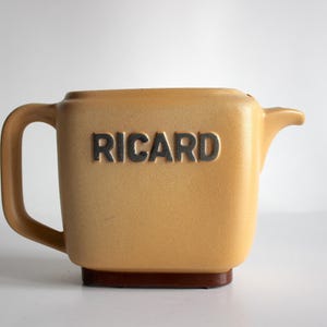 Puede incluir: Una jarra de cerámica de color beige con una base marrón y un asa. La jarra tiene la palabra "RICARD" en relieve en la parte delantera.
